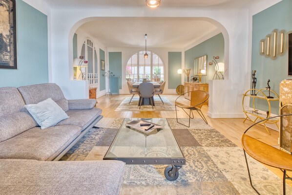 A spacious living room with a sofa, coffee table, and chairs, an archway leading to a dining area, and a view through the archway.
