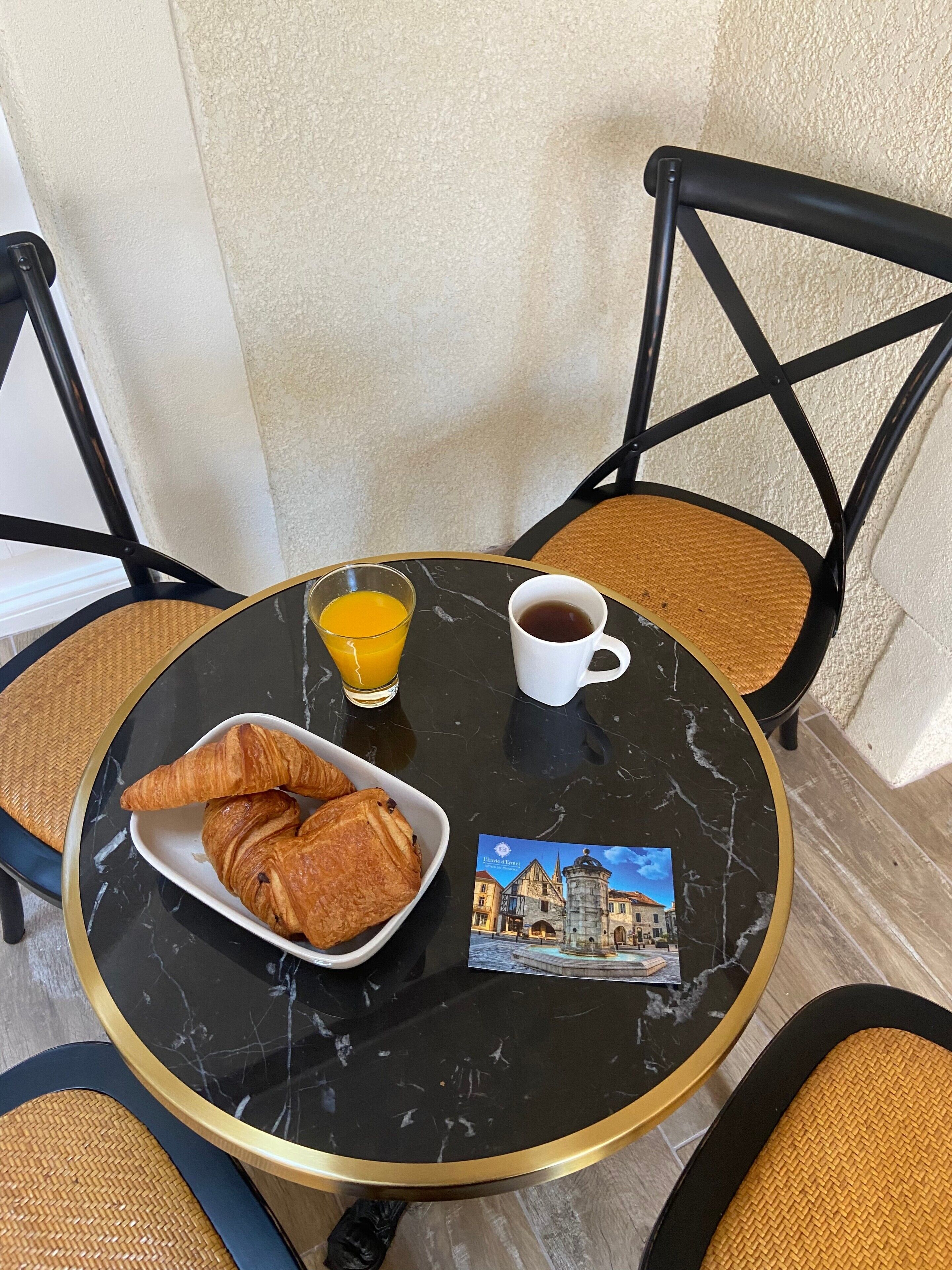 Une table ronde avec un plateau en marbre noir, une assiette blanche contenant des croissants, un verre de jus d’orange, une tasse de café blanche et une carte postale.