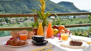 Une table de petit-déjeuner avec des croissants, du jus de fruits et des viennoiseries, donnant sur un paysage pittoresque avec des collines et une étendue d’eau.