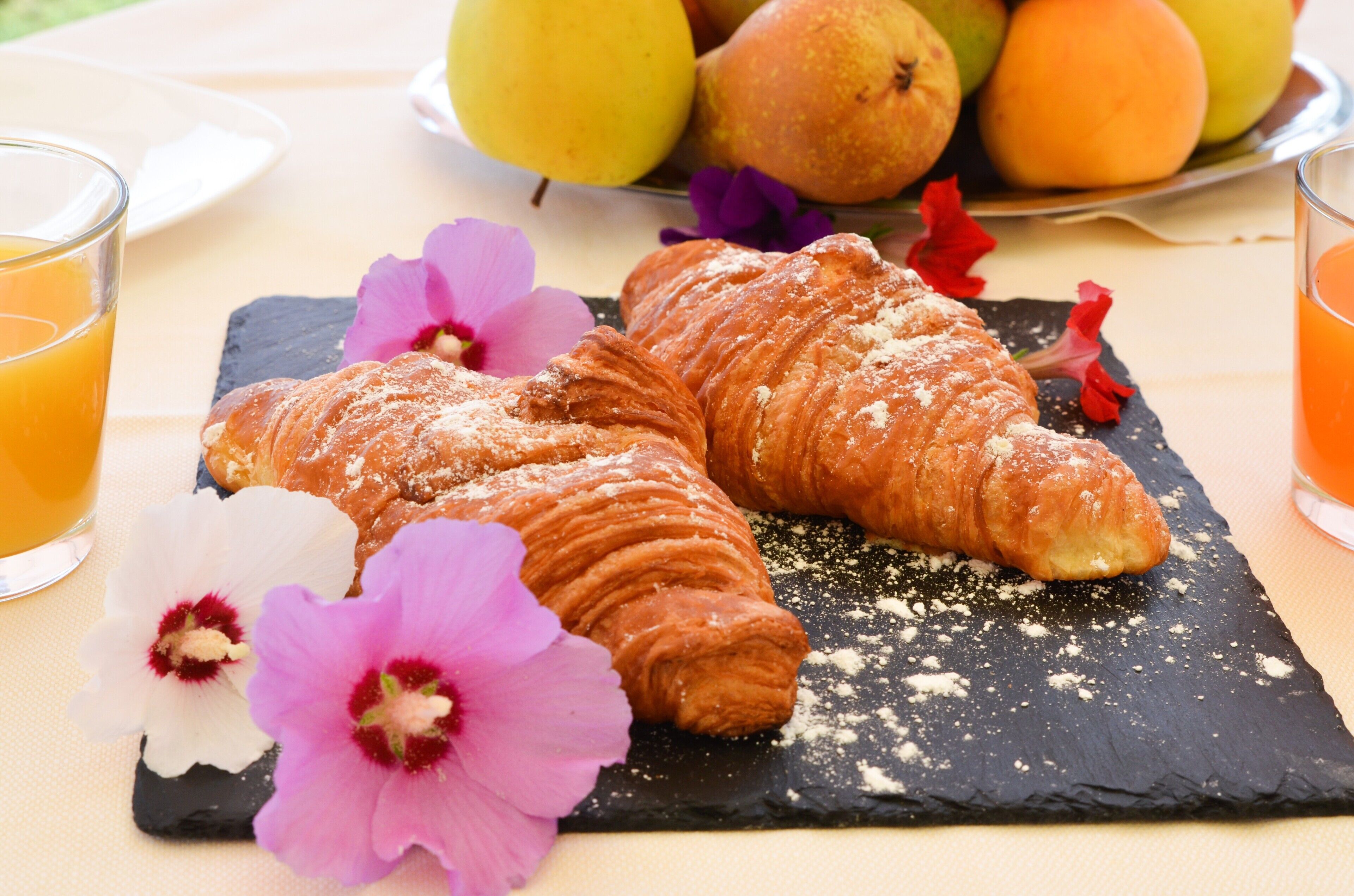 A breakfast setting with croissants on a slate board, accompanied by flowers and a fruit plate.