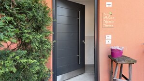 Una puerta negra con manija, un taburete de madera pequeño con un cubo morado y un cartel con nombres de habitaciones.