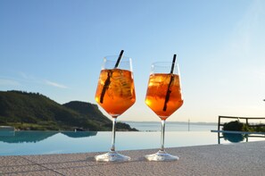 Two glasses of amber-colored beverage with straws on a terrace overlooking a calm body of water and a hilly landscape.