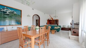 Uma sala de jantar com mesa e cadeiras de madeira, um quadro na parede e um sofá verde ao fundo.