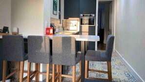 Een compacte keuken met een inbouwoven, een klein eetgedeelte met vier grijze stoelen, en een betegelde vloer.