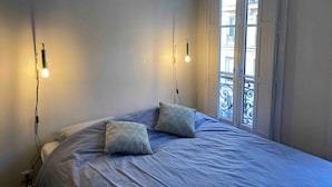 Een slaapkamer met een bed, twee kussens met een patroon, een nachtkastje, een wandlamp en een raam met uitzicht op gebouwen.