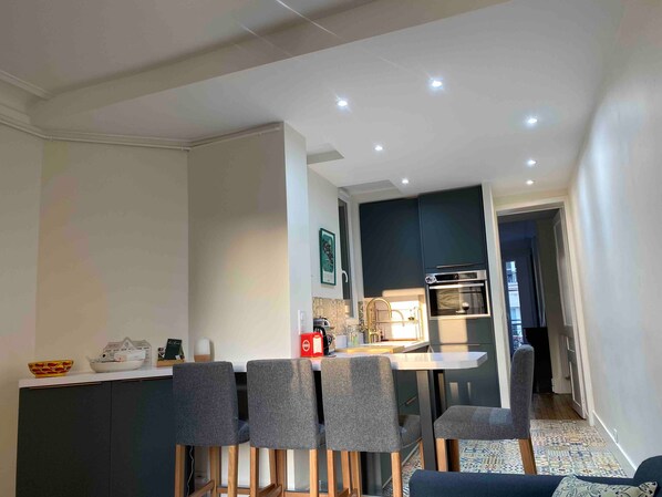 A modern kitchen with dark cabinets, a white countertop, and a built-in oven. There are four grey chairs around a table with a small decorative bowl on it.