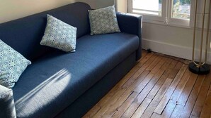 Een kamer met een donkerblauwe bank, gekleurde kussens, een houten vloer, een groot raam met uitzicht en een hoge staande lamp.