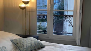 Een slaapkamer met een bed, een raam met uitzicht op gebouwen en een vloerlamp.