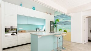 A modern kitchen with white and light blue cabinets, a central island with stools, a refrigerator, and a mural of a beach scene. - Architect's Villa 800m from the Sea – Comfort, Nature, and Serenity in Oléron, with Terrace Garden (Dolus-d'Oleron)