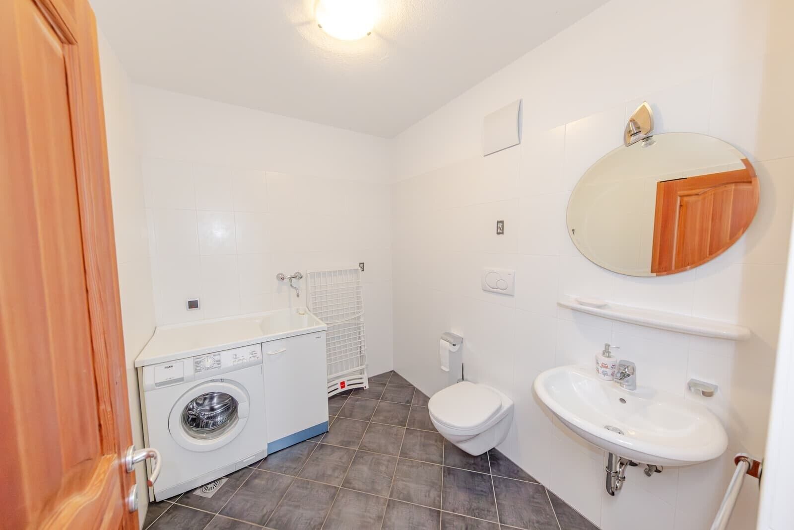 A compact bathroom with a washing machine, toilet, sink, and a mirror with a wooden cabinet.