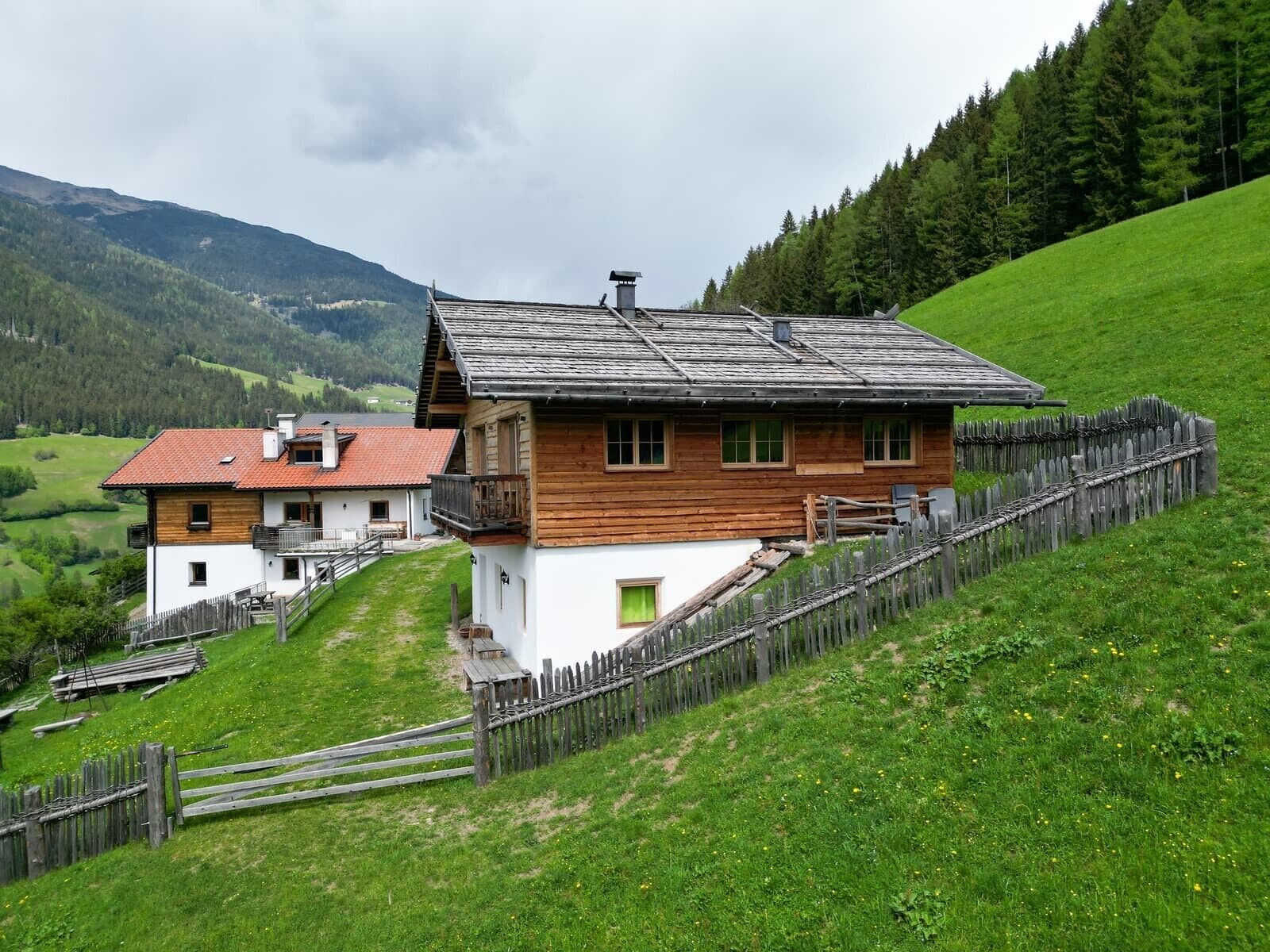 A wooden house with a balcony, surrounded by a wooden fence and a grassy hill.