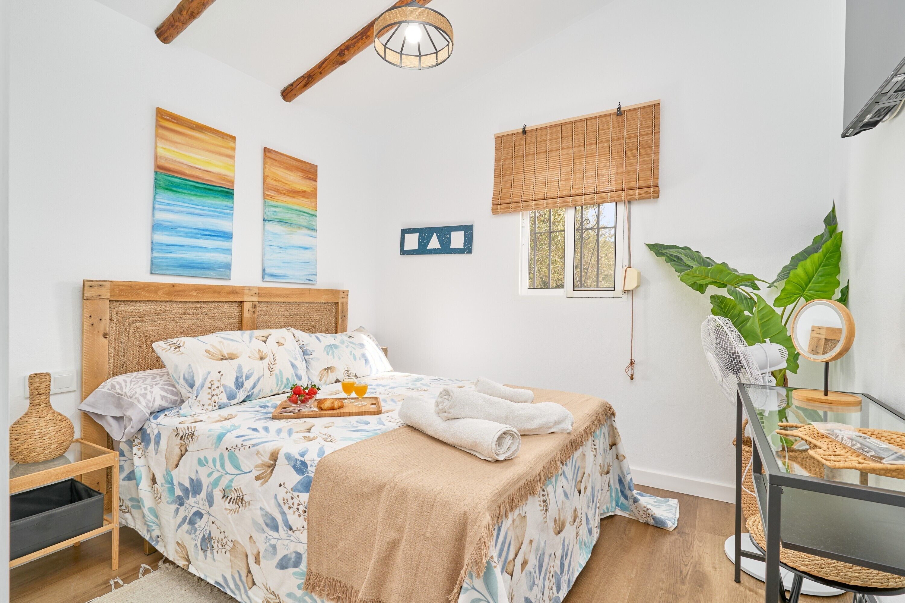 Une chambre avec un lit, une table de chevet, un ventilateur fixé au mur, une fenêtre donnant sur un beau paysage et une grande plante.