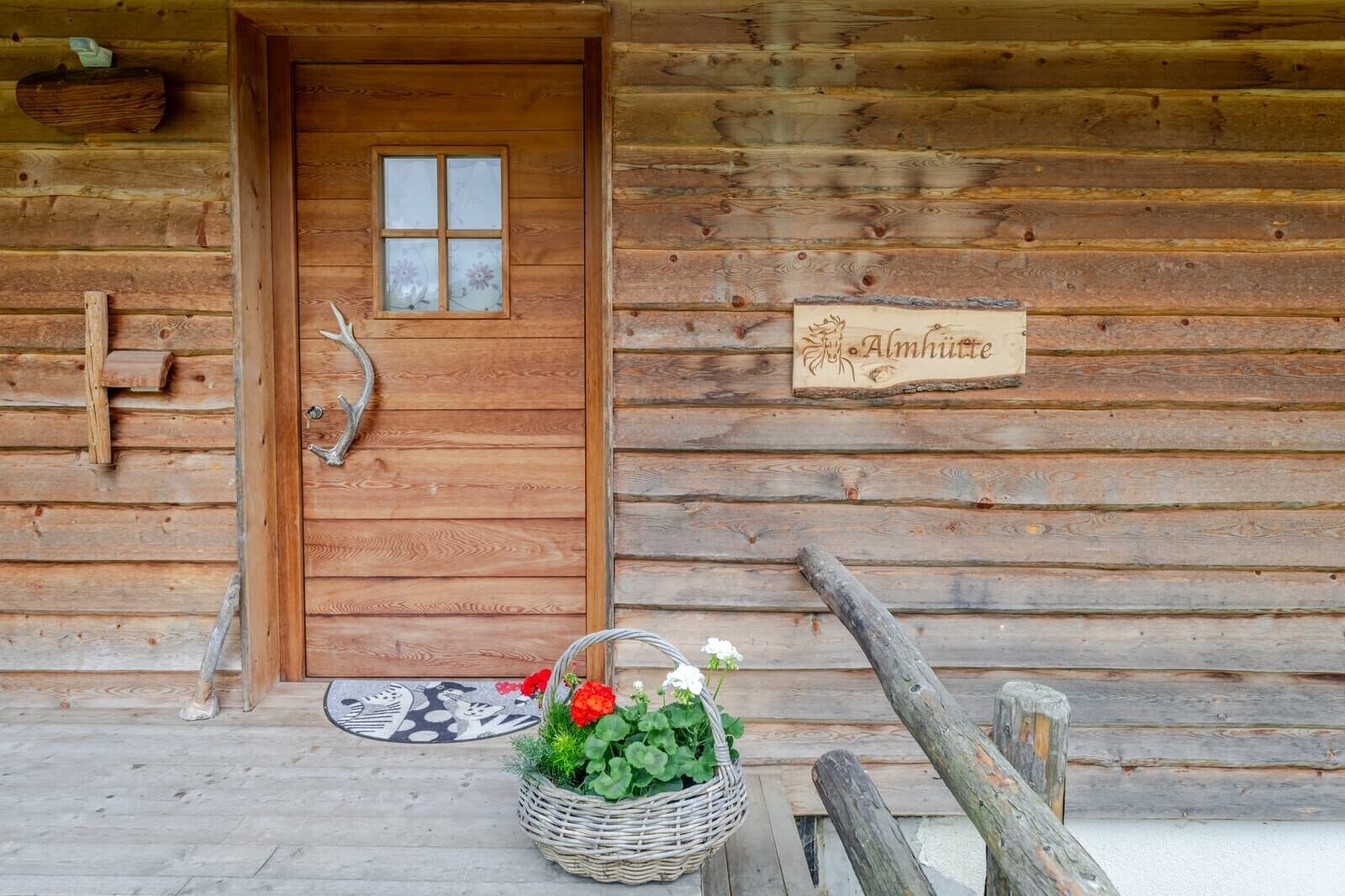 Eine Holzhütte mit Holztür, einem Fenster mit Frostglas, einem Schild mit dem Namen 'Almhütte' und einem Korbwägelchen mit Blumen auf der Veranda.