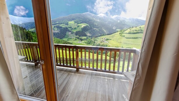 Ein Holzbalkon mit Glastür, der auf eine gebirgige Landschaft mit grünen Feldern und klarem Himmel blickt.