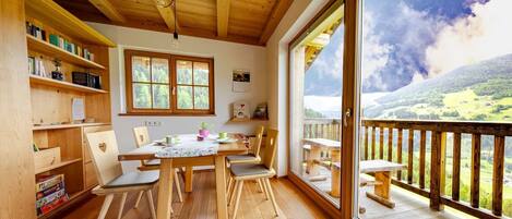 A wooden interior with a dining table, chairs, a bookshelf, and a large window with a mountain view.