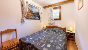 Une chambre à coucher avec un lit, une chaise en bois, une table de chevet avec une lampe, une fenêtre avec des rideaux et un tableau accroché au mur.