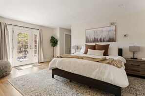 Un dormitorio con una cama grande, una mesita de noche, una lámpara, un cuadro y un ventanal corredero que da a un espacio exterior.