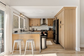 Una cocina moderna con armarios de madera, una isla central con taburetes, un refrigerador de acero inoxidable y una ventana con persianas.