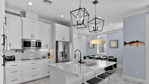 Una cocina moderna con armarios blancos, una isla central con taburetes de bar, electrodomésticos de acero inoxidable y una lámpara colgante.