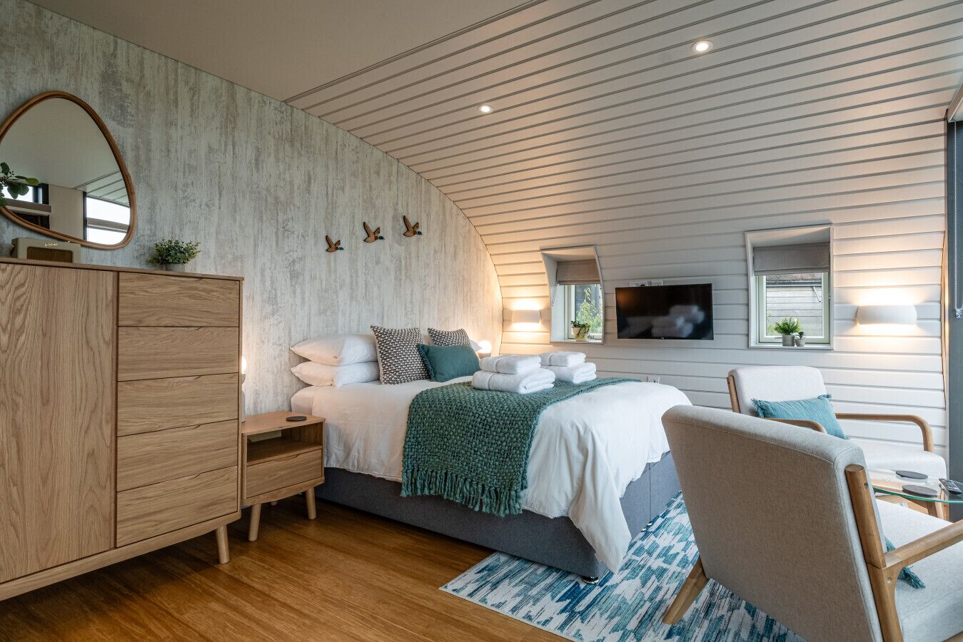 A modern bedroom with a wooden dresser, a bed with white linens and a green throw, a chair, and a TV mounted on the wall.