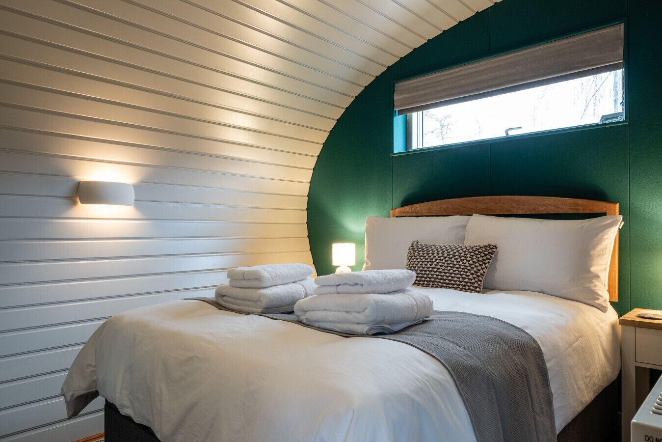A neatly made bed with white linens and a grey throw blanket, a bedside lamp, and a window with a brown blind.