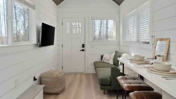 A modern, compact kitchen with white wooden walls, a built-in TV, and a dining area with a bench and table set for four. - Tiny Home · Hot Tub · Fire Pit · Romantic Escape (Laurelville)