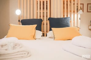 A hotel room with two beds, each with yellow pillows, a wooden partition, and a round table with a lamp.