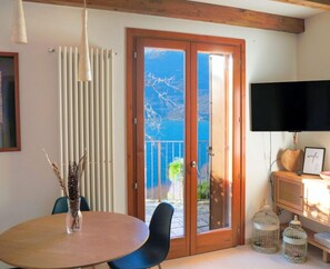 A room with a wooden ceiling, a round table with chairs, a TV on the wall, and a sliding glass door leading to an outdoor area with a view of the sea.