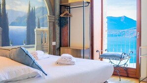 A bed with white linens, a view of the sea through a balcony door, and a painting of a landscape on the wall.