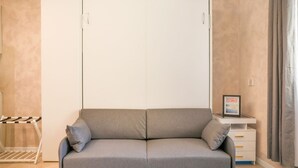 A room with a grey sofa, a white wardrobe, a small white side table with a picture frame, and a window with curtains. - BLUE WAVE by Design Studio (Bellano)
