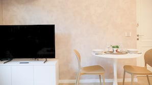 A modern living room with a flat-screen TV on a white cabinet, a round dining table with four chairs, and a door in the background. - BLUE WAVE by Design Studio (Bellano)