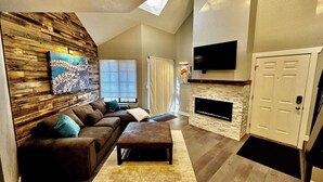 A modern living room with a brown leather sofa, a matching ottoman, and a large abstract painting on the wall. A flat-screen TV is mounted above a stone fireplace. - ~Remodeled Mtn View Condo, Hot Tub, Modern, Fast WiFi~ (Boulder)