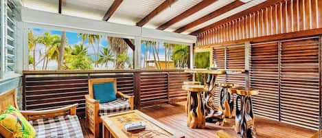 A covered outdoor seating area with wooden furniture, a ceiling fan, and a view of palm trees through the window.
