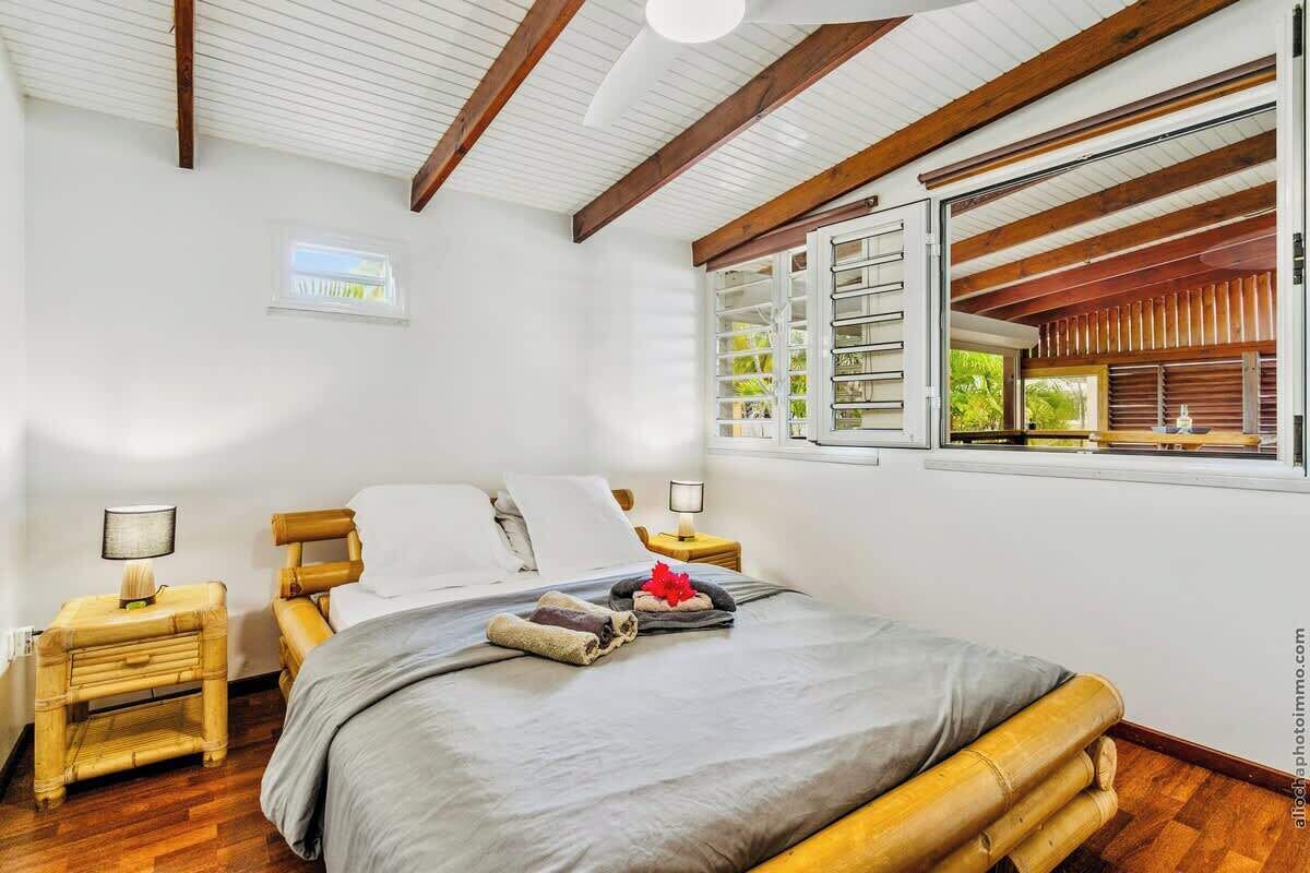 A bedroom with a bed, wooden headboard, two bedside tables with lamps, a window with blinds, and a view of greenery outside.