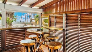 Eine Holzterrasse mit Tisch und Hocker, mit Blick auf einen Strand mit Palmen und einem klaren Himmel.