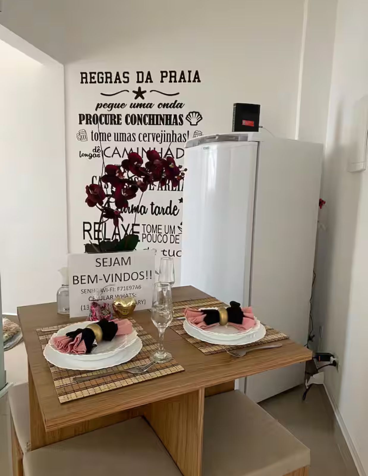 Een eethoek met een houten tafel gedekt voor twee personen, een decoratieve wand met tekst en bloemen, en een witte koelkast op de achtergrond.
