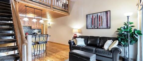 A spacious living area with a wooden staircase, a black leather sofa, a dining area, and a large painting on the wall.