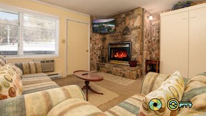 Ein Wohnzimmer mit Kamin aus Stein, einem an der Wand befestigten Fernseher und einem großen Fenster mit Jalousien.