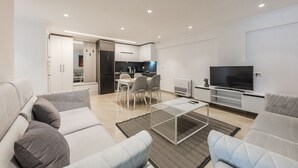 A modern living room with a sofa, a coffee table, and a TV. There is a dining area with chairs and a table. The kitchen is visible in the background.