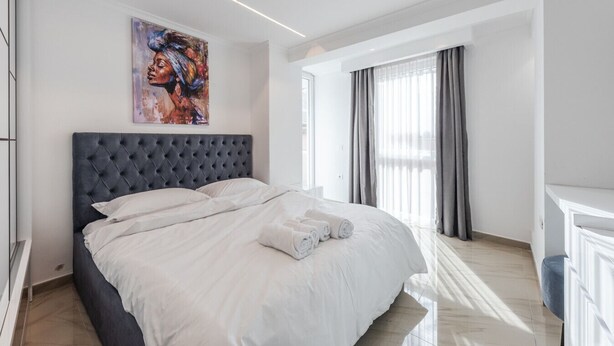 A modern bedroom with a large bed, a tufted headboard, a painting on the wall, and a window with curtains.