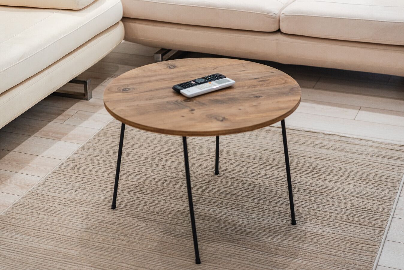 Ein runder Holztisch mit einer Fernbedienung darauf, auf einem strukturierten Teppich in einem Wohnzimmer mit einer beigen Couch.