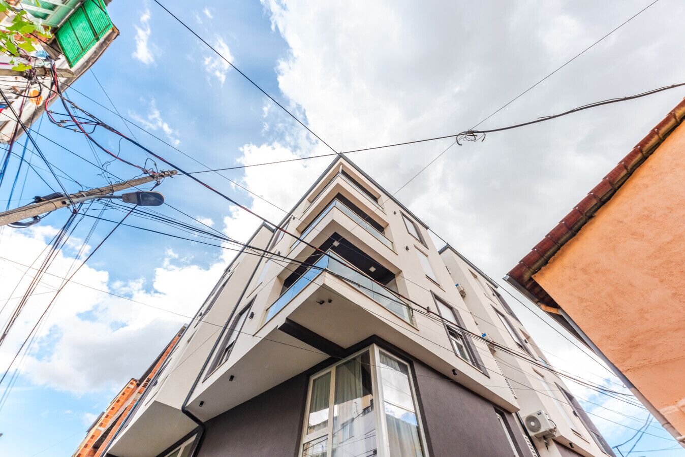 A modern building with a prominent corner balcony, multiple windows, and visible power lines against a partly cloudy sky.