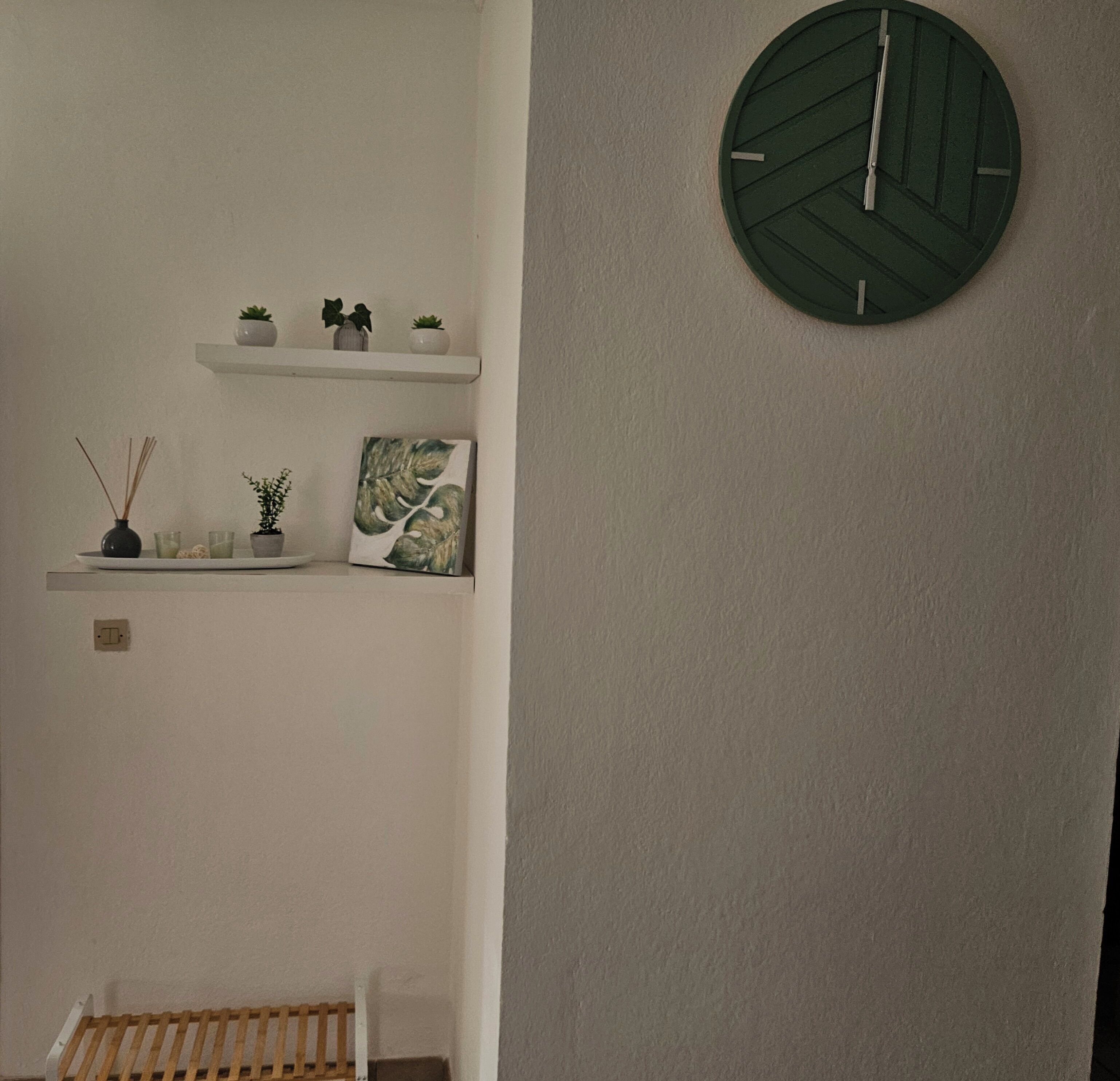 A room with a white wall, a round green clock, a white shelf with plants and a picture, and a wooden crib.