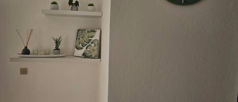 A room with a white wall, a round green clock, a white shelf with plants and a picture, and a wooden crib.