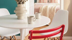 A room with a white table, a red chair, a teal wall, and a bed with striped pillows.