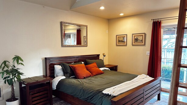 Un dormitorio ordenado con una cama de madera, una mesita de noche y una planta en maceta, además de una ventana con cortinas.