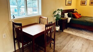 Una habitación con una mesa de comedor y sillas, una cama con almohadas, una mesita de noche con lámpara, y una ventana con vista a árboles.