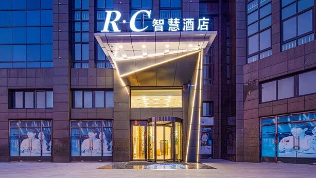 A hotel entrance with a modern design, featuring a prominent 'R.C' sign above the entrance and a glass door leading inside. - RC SMART HOTEL (Yancheng)