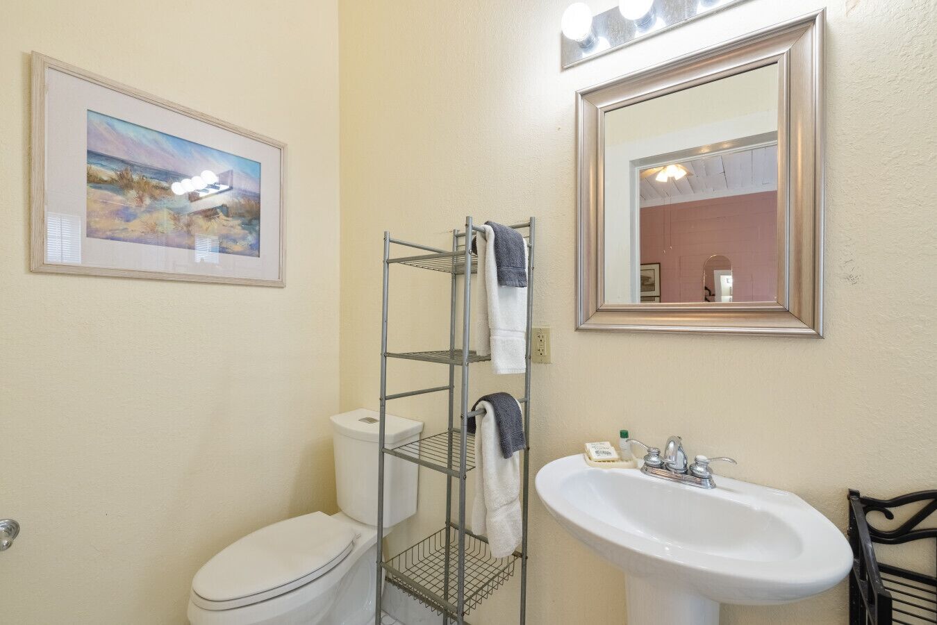 Een badkamer met een toilet, een wastafel, een metalen plank met handdoeken, een ingelijst plaatje en een spiegel.