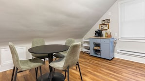 Un coin repas avec une table ronde et quatre chaises, un petit meuble contenant des ustensiles de cuisine et une fenêtre avec des stores.